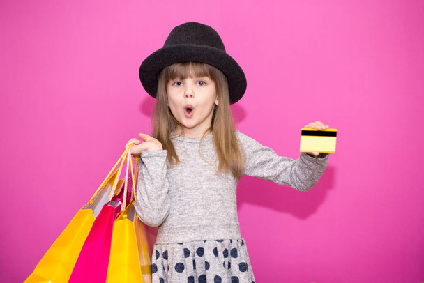 Little girl with shopping bags and shocked face hold credit card isolated on pink background - Photo, Image