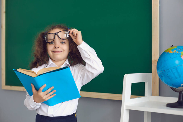 Back to school.Smart schoolgirl in glasses with a book in her hands on the background of the school blackboard - Φωτογραφία, εικόνα