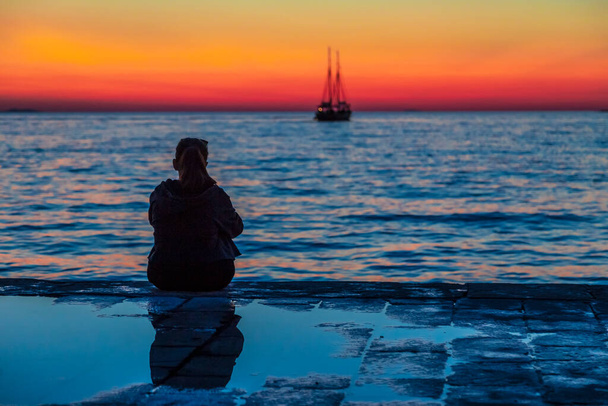 夕暮れ時に海を眺める若い女性のシルエット。アドリア海｜ヨーロッパ・クロアチア・ザダル町. - 写真・画像