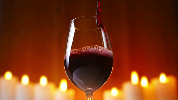 czerwone wino wlewające się do kieliszka z płonącymi świecami na tle - Materiał filmowy, wideo
