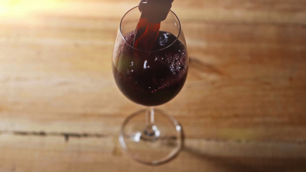 powolny ruch czerwonego wina wlewającego się do kieliszka na drewnianym stole - Materiał filmowy, wideo