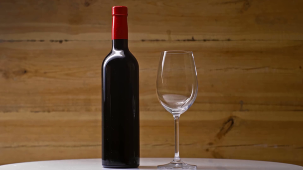 bottle of red wine near empty wineglass spinning on wooden background - Filmati, video
