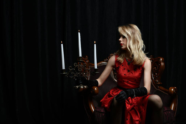 красива дівчина в червоній сукні позує в кріслі з канделябром на чорному тлі
 - Фото, зображення