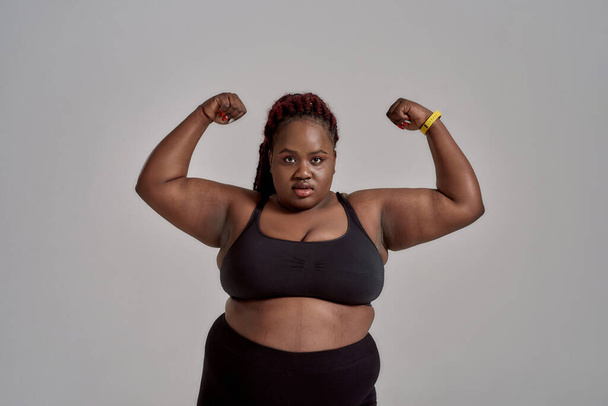 Plump, plus size african american woman in black sportswear looking at camera, showing strong arms in studio over grey background. Concept of sport, healthy lifestyle, body positive, equality - Photo, image