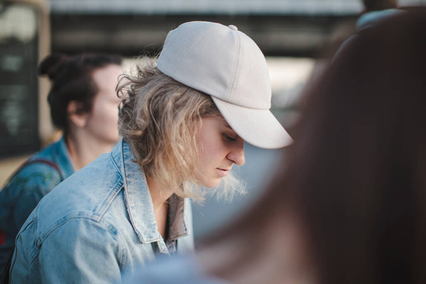 Beautiful blonde girl in a cap with short hair sits in a group of girls, looks down. Selective focus - Foto, Bild