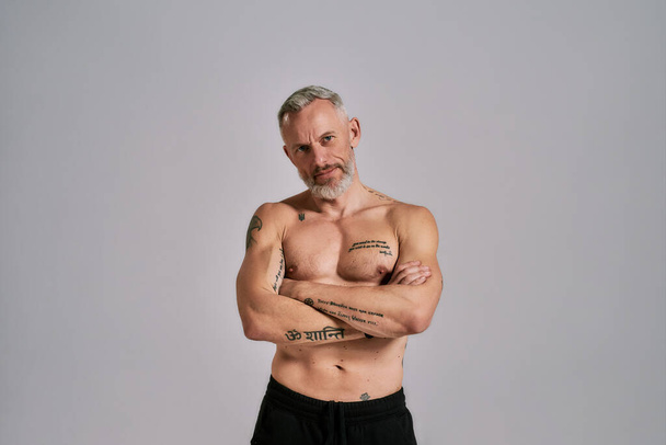Be an inspiration. Half naked middle aged muscular man looking at camera, showing his body, while posing in studio over grey background - Photo, image