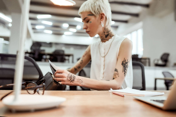 Looking for sales report. Young blonde tattooed businesswoman with short haircut trying to find something in her handbag while sitting at her desk in office - Photo, Image
