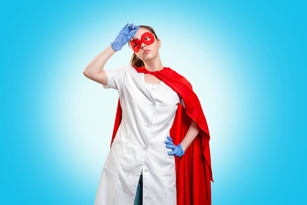 A tired doctor in a mask, medical gloves, and red superhero Cape wipes sweat from her forehead. Blue background. The concept of the Power of a super hero for medicine and stress at work. Copy space. - Foto, immagini