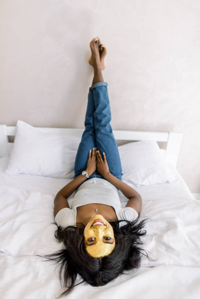 Young pretty African American woman with golden peel off facial mask resting, lying on bed, enjoying facial spa treatment at home. Skin care, beauty and spa at home - Photo, Image