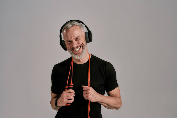 Middle aged muscular man in black t shirt and headphones smiling at camera holding jump rope, posing in studio over grey background - Foto, immagini