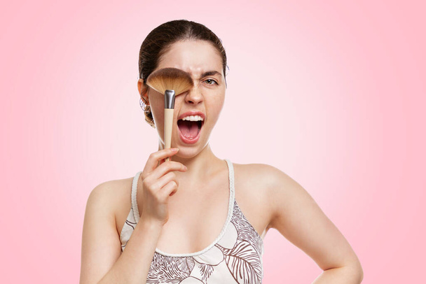 A young pretty woman covers one eye with a makeup brush and screams. Pink background. Copy space. The concept of makeup, self-care and cosmetics. - 写真・画像