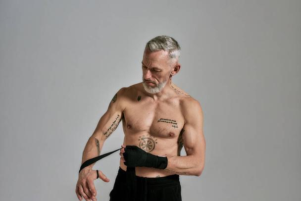 Half naked middle aged athletic man, kickboxer wrapping hands for Muay Thai, Boxing or Kickboxing, standing in studio over grey background - Photo, Image
