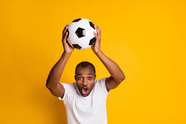 Photo of attractive dark skin guy man hold leather football ball above head addicted fan screaming supporter wear casual white t-shirt isolated yellow vibrant color background - Zdjęcie, obraz