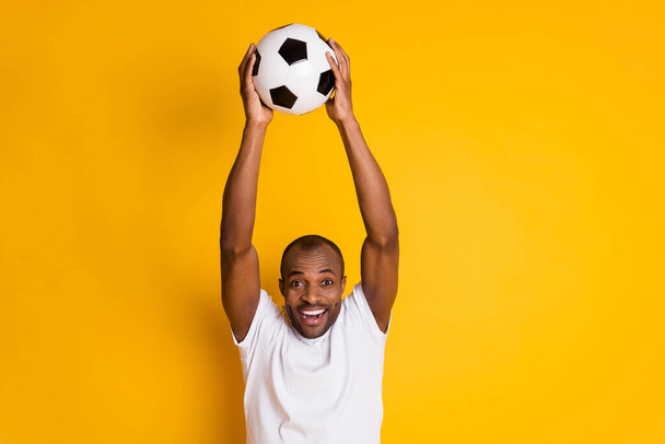 Delighted positive cheerful afro american guy hold football ball celebrate national team goal world cup match wear white t-shirt isolated over bright shine yellow color background - Φωτογραφία, εικόνα
