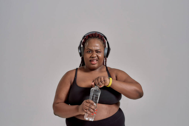 Plump, plus size african american woman in sportswear and headphones smiling, opening bottle of water, posing in studio over grey background - Foto, afbeelding