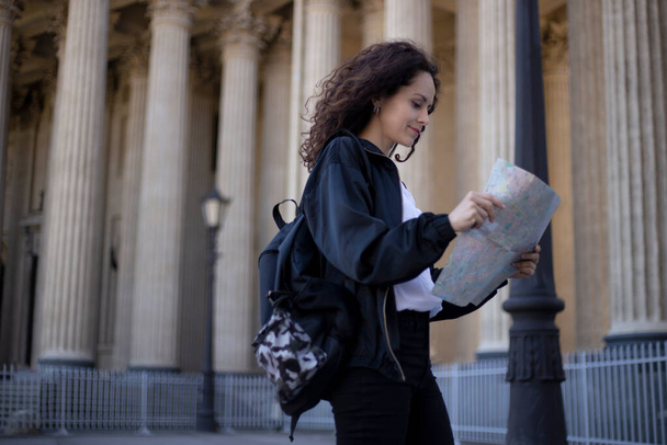 Portrait of a young woman with a city map in hands, looking on a historic city, have a backpack, over cathedral background. - Photo, Image
