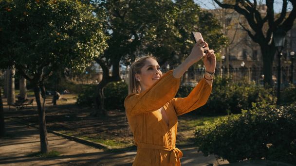 Joven hermosa mujer elegante tomando fotos en el teléfono inteligente caminando en el parque de la ciudad. Elegante dama tomando selfie al aire libre
 - Foto, imagen