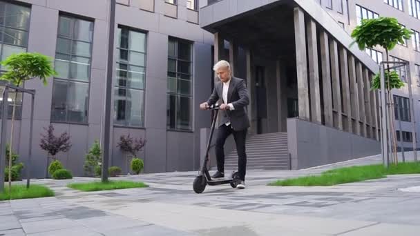 Attractive satisfied stylish young light-haired guy enjoying his ride on own e-scooter near beautiful architectural building - Filmagem, Vídeo