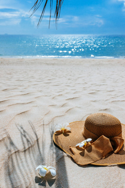 Straw hat with frangipani flowers on white sea sand. With beautiful seascape, the blue sea in the background, Palm leaf shadow. Copy space, background for advertisement of a seaside vacation - Fotó, kép