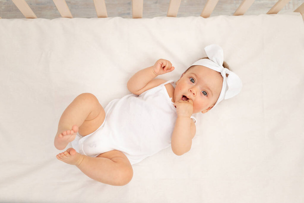 bébé fille de 6 mois avec les doigts dans la bouche couché dans un berceau dans une chambre d'enfants avec des vêtements blancs sur le dos et en regardant la caméra, le matin de bébé, concept de produits pour bébé. - Photo, image