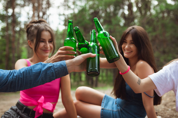 Group of friends clinking beer bottles during picnic in summer forest. Lifestyle, friendship - Zdjęcie, obraz