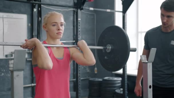 Slow motion of young woman doing barbell squats training with instructor in gym - Video