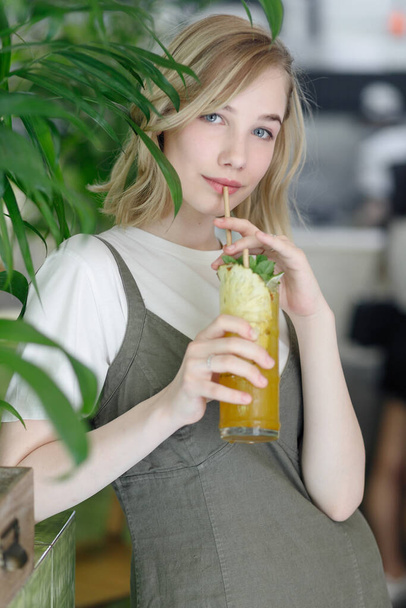 Close up young cheerful woman in dress happily looking in camera with big green leaves on background in cozy green cafe. Girl in glasses. holding a drink in his hands - Foto, imagen