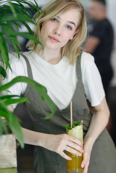 Close up young cheerful woman in dress happily looking in camera with big green leaves on background in cozy green cafe. Girl in glasses. holding a drink in his hands - Фото, изображение