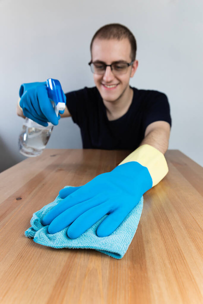 Young man cleaning a wooden table with blue cloth and alcohol sanitizer, wearing blue gloves, glasses and smiling. Vertical. Coronavirus disinfection in the house - Photo, Image