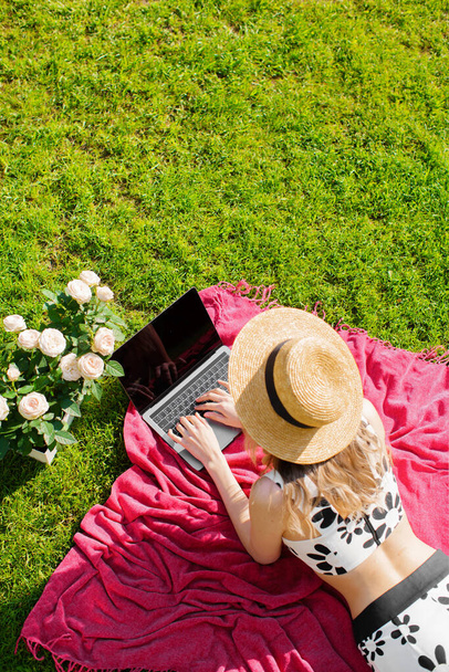 Young girl blogger in a hat working on a laptop with empty screen on green lawn, photo for a blog or ad - Foto, immagini