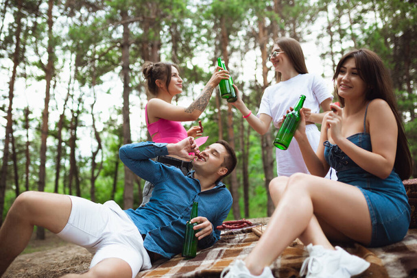 Grupo de amigos batendo garrafas de cerveja durante piquenique na floresta de verão. Estilo de vida, amizade
 - Foto, Imagem