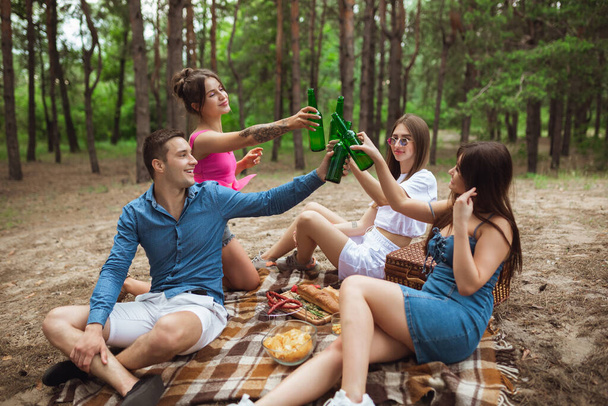 Group of friends clinking beer bottles during picnic in summer forest. Lifestyle, friendship - 写真・画像