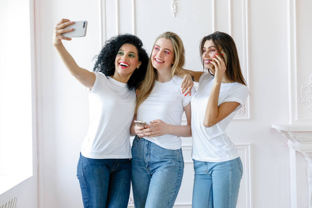 Portrait of three seductive multiethnic women standing together and smiling. Three beautiful smiling girlfriends taking selfie with mobile phone. taking self portrait. - Valokuva, kuva