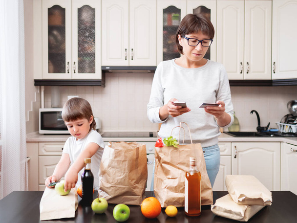 Woman and toddler boys sorts out purchases in the kitchen. Kid bites an apple. Grocery delivery in paper bags. Subscription service from grocery store. Mother and son at kitchen. - Foto, Bild