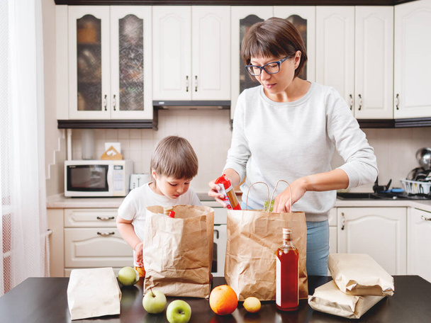 Woman and toddler boy sorts out purchases in the kitchen. Kid bites an apple. Grocery delivery in paper bags. Subscription service from grocery store. Mother and son at kitchen. - Foto, Bild