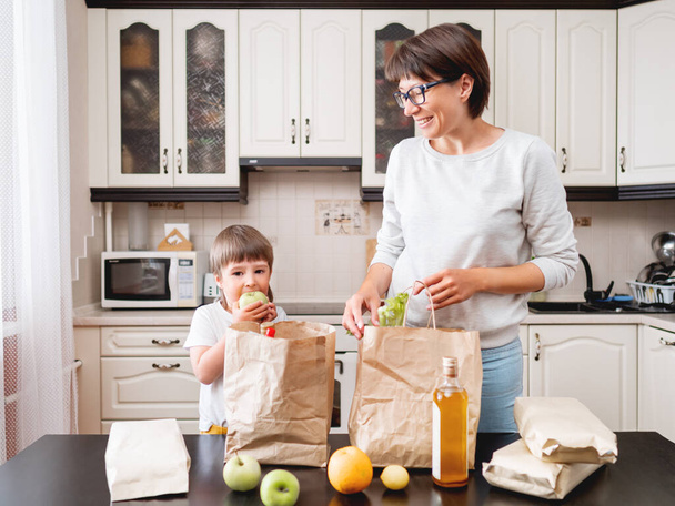 Woman and toddler sorts out purchases in the kitchen. Kid bites an apple. Grocery delivery in paper bags. Subscription service from grocery store. Mother and son at kitchen. - Foto, Bild