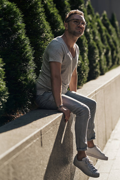 stylish guy with glasses of seats on a concrete handrail near green plants, looks at the top and thinks. light jeans and a fashionable gray T-shirt. beautiful contour sunlight. - Photo, Image