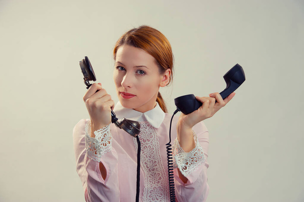 Sercretary overwhelmed. Serious woman holding, giving two telephones and looking seriously at camera. Caucasian Business person, pink formal shirt, redhead ginger hair isolated on light grey studio background - Foto, imagen