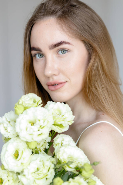 Cute happy young girl in white blank t-shirt, holding a bouquet and looks at the of flowers, enjoying the smell. beautiful blond teen girl with braces on her teeth smiling - Фото, зображення