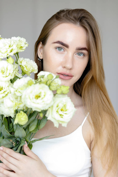 Cute happy young girl in white blank t-shirt, holding a bouquet and looks at the of flowers, enjoying the smell. beautiful blond teen girl with braces on her teeth smiling - Фото, изображение