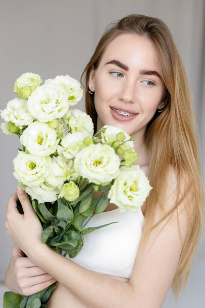 Cute happy young girl in white blank t-shirt, trzymając bukiet i patrzy na kwiaty, ciesząc się zapachem. piękne blondynka nastolatek dziewczyna z aparat na jej zęby uśmiech - Zdjęcie, obraz