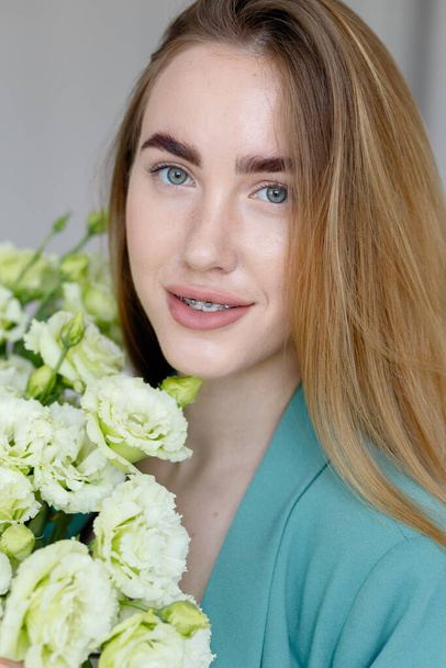 Cute happy young girl in white blank t-shirt, holding a bouquet and looks at the of flowers, enjoying the smell. beautiful blond teen girl with braces on her teeth smiling - Foto, Imagen