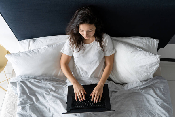 Beautiful brunette girl in bed with a laptop. Work from morning to late night. Electronic gadgets before bedtime or in the morning - Fotoğraf, Görsel