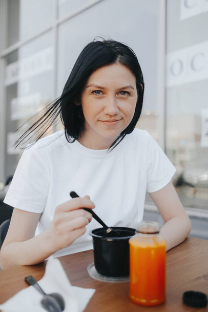 A young woman with black hair in a white T-shirt sits on an open veranda in a cafe and eats food from disposable plastic dishes.  - Foto, afbeelding
