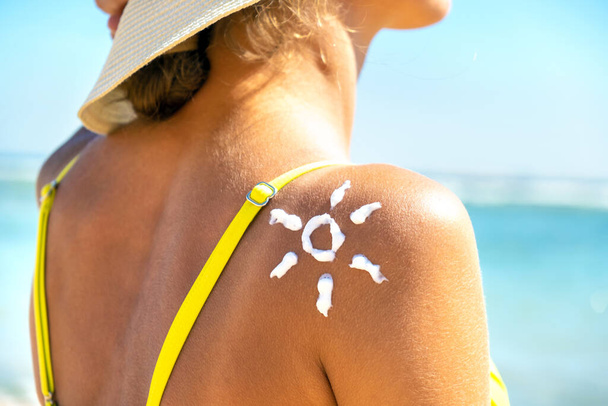 Achteraanzicht van een jonge bruinende vrouw op het strand met zonnecrème in de vorm van een zonnebrandcrème op haar schouder. UV zonnebrandbescherming en zonnecrème huidverzorging concept - Foto, afbeelding