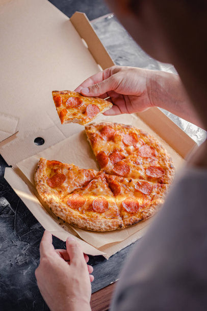 Мужская рука держит кусочек пиццы из коробки с доставкой. Концепция быстрого питания
. - Фото, изображение