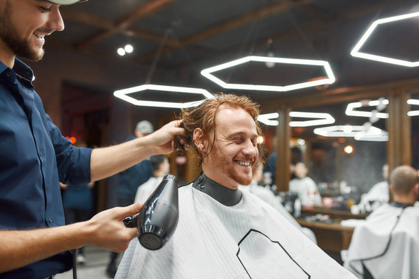 Great day in barbershop. Side view of young smiling redhead guy sitting in barbershop chair while barber drying his wet hair - Foto, imagen