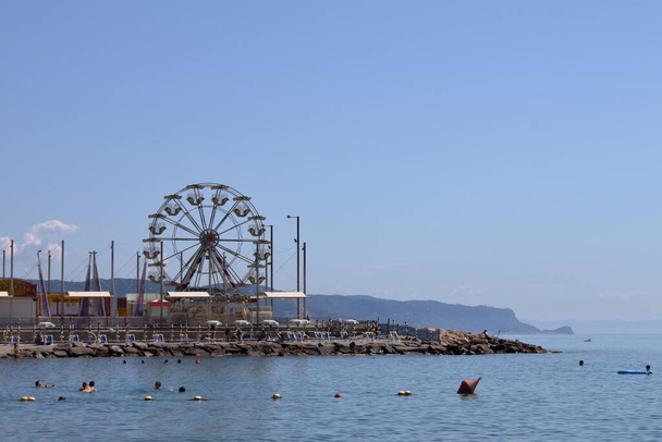 Liguria, Włochy - lato 2020: turyści kąpią się w zatoce Liguryjskiej, w tle diabelski młyn na szczycie molo - Zdjęcie, obraz