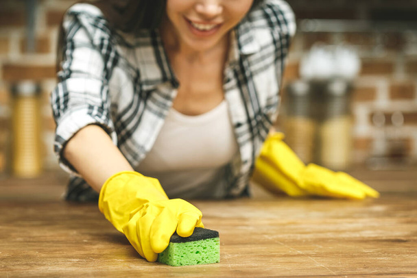 Belle femme dans la cuisine sourit et essuie la poussière à l'aide d'un spray et d'un plumeau pendant le nettoyage de sa maison, gros plan - Photo, image