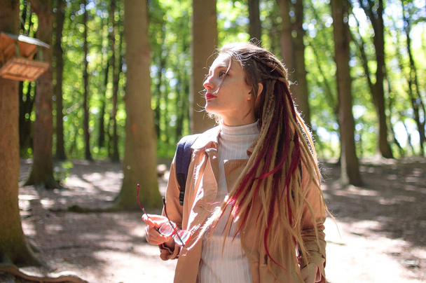 Young woman wearing glasses, casual nude trench, and dreadlocks is standing at the urban park on trees background during sunny spring day outdoors - Photo, Image